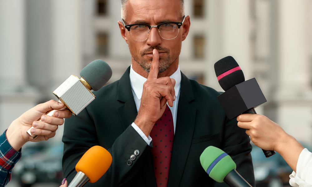 Man in suit shushing many microphones from journalists