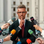Man in suit shushing many microphones from journalists
