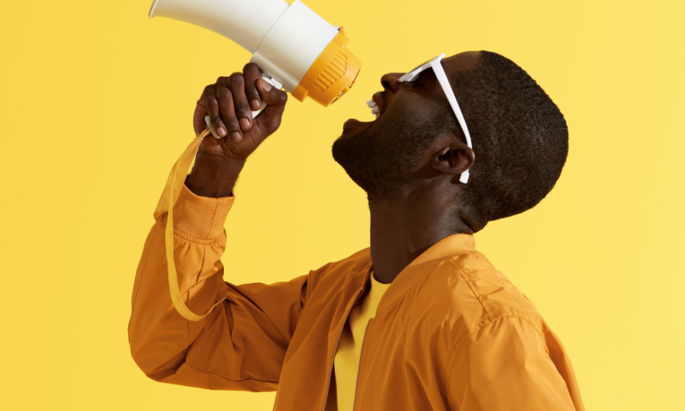 A man shouting into a megaphone in front of a yellow background